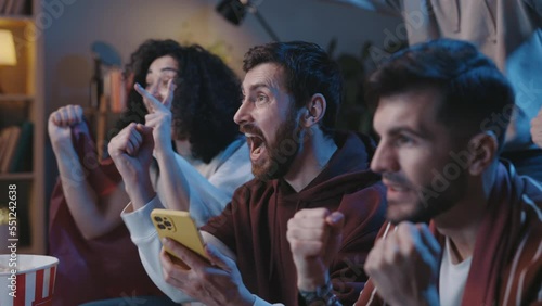 Excited soccer fans sitting at home watching game on tv, using smartphone app to online bet. Friends celebrating victory during football game.