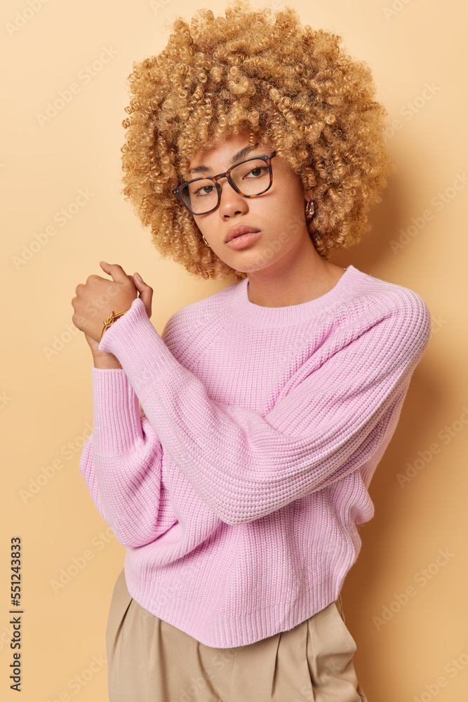 Self confident woman looks seriously at camera keeps hands together has determined assured expression wears eyeglasses long sleeved pink jumper and trousers stands indoors against beige background Self confident woman looks seriously at camera keeps hands together has determined assured expression wears eyeglasses long sleeved pink jumper and trousers stands indoors against beige background