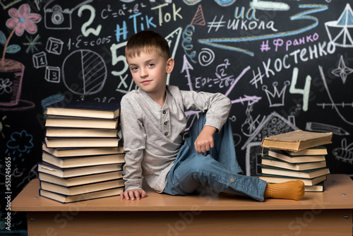 a little boy of Slavic European appearance sits on a desk near a pile of books against a slate wall