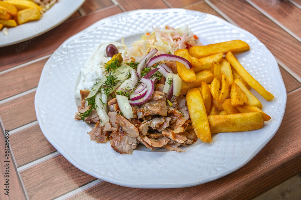 Gyros, Greek dish from sliced meat roasted on a turning spit, served with French fries, coleslaw, tzatziki and onions on a white plate, wooden outdoor table, selected focus