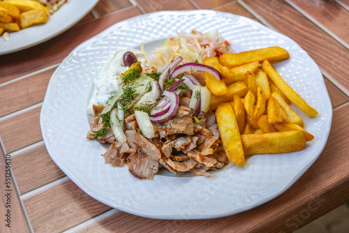 Gyros, Greek dish from sliced meat roasted on a turning spit, served with French fries, coleslaw, tzatziki and onions on a white plate, wooden outdoor table, selected focus