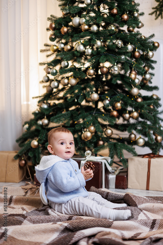 A 2-year-old little boy sits near a Christmas tree decorated with toys. New Year's atmosphere
