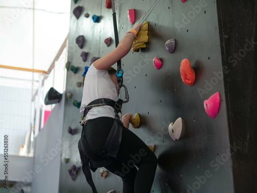 A little girl trains in mountaineering, climbs a wall with obstacles in a protective holding belt in a children's amusement park, climbs a climbing wall