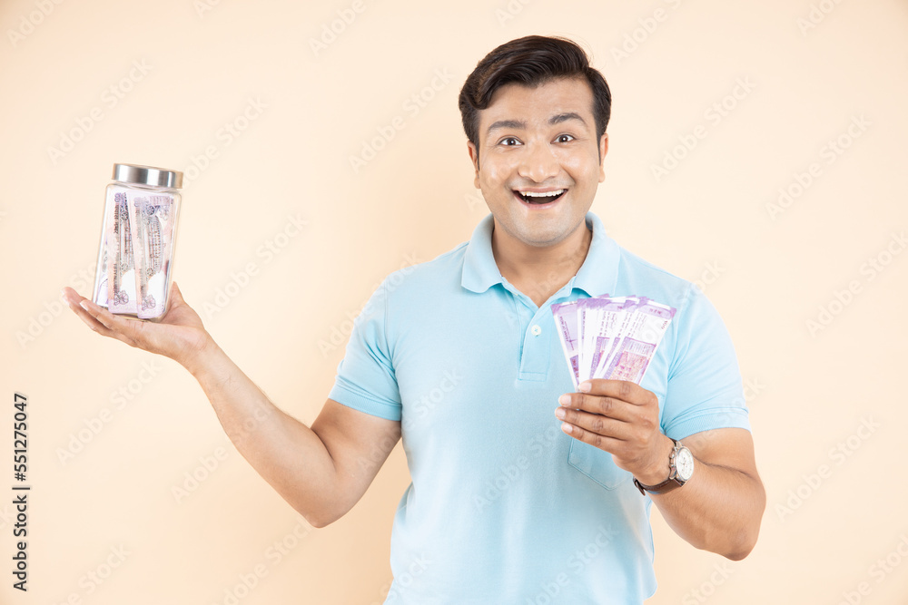Amazed or cheerful handsome man with Indian rupee notes in one hand and glass jar with lots of cash money in other hand isolated on beige studio background. saving and smart investing concept. Amazed or cheerful handsome man with Indian rupee notes in one hand and glass jar with lots of cash money in other hand isolated on beige studio background. saving and smart investing concept.