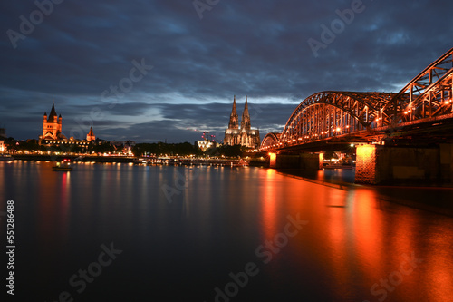 Cologne, Germany. Famous Hohenzollern Bridge over Rhine river. Buildings in historic city centre. Towers of Cathedral and city hall by night. 