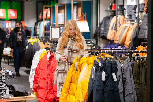 Young female customer with golden hair choosing clothes in modern clothing store.