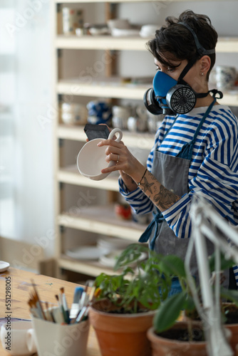 Young female artisan concentrated on modelling mug from clay and polishes the finished product in a respirator in creative studio. Woman ceramic business owner making craft for sale