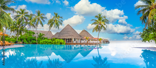 Beachside tourism panoramic landscape. Luxury beach resort poolside  swimming pool and beach chairs beds umbrellas with tropical palm trees and blue sky. Summer travel and vacation background panorama