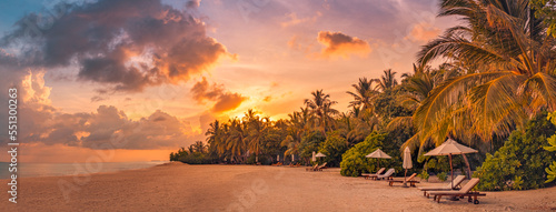 Fantastic panoramic view. Sandy shore with orange sunrise sunset sunlight over chairs palm trees. Tropical island beach landscape, exotic coast. Summer vacation, holiday amazing nature. Relax panorama