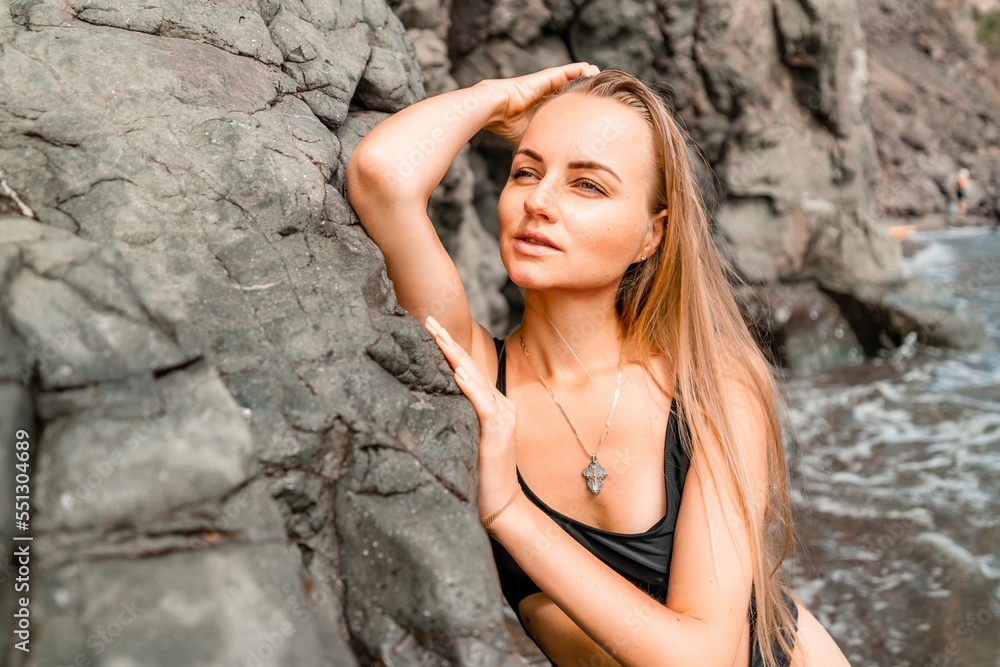Woman swimsuit sea. Attractive blonde woman in a black swimsuit enjoying the sea air on the seashore around the rocks. Travel and vacation concept.