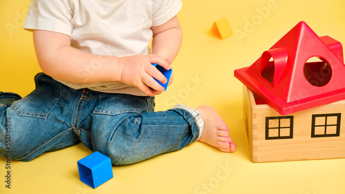 Toddler baby is playing sorter logic educational games with geometric shapes on a studio yellow background. Happy child play with educational toy house, learning logic. Kid aged one year four months