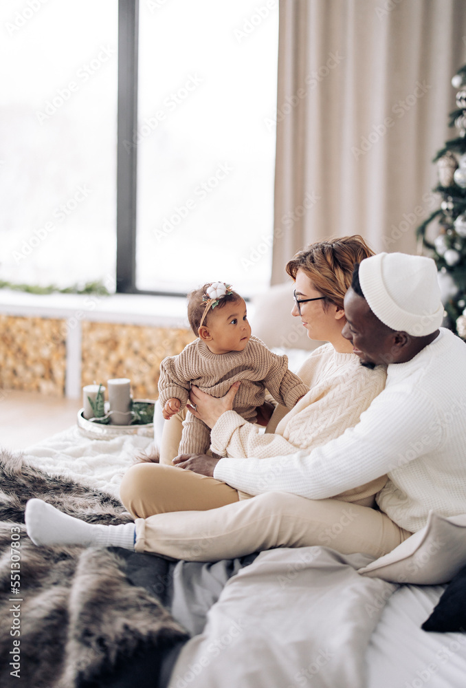 Happy interracial family sits on bed and plays with their baby daughter against background of Christmas tree and window. Happy interracial family sits on bed and plays with their baby daughter against background of Christmas tree and window.