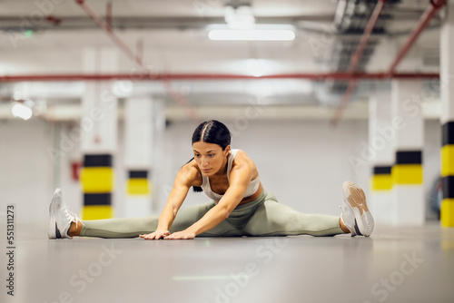 A flexible sportswoman is sitting on garage floor and stretching her legs.