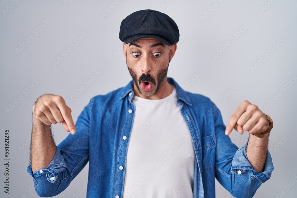 Hispanic man with beard standing over isolated background pointing down with fingers showing advertisement, surprised face and open mouth Hispanic man with beard standing over isolated background pointing down with fingers showing advertisement, surprised face and open mouth