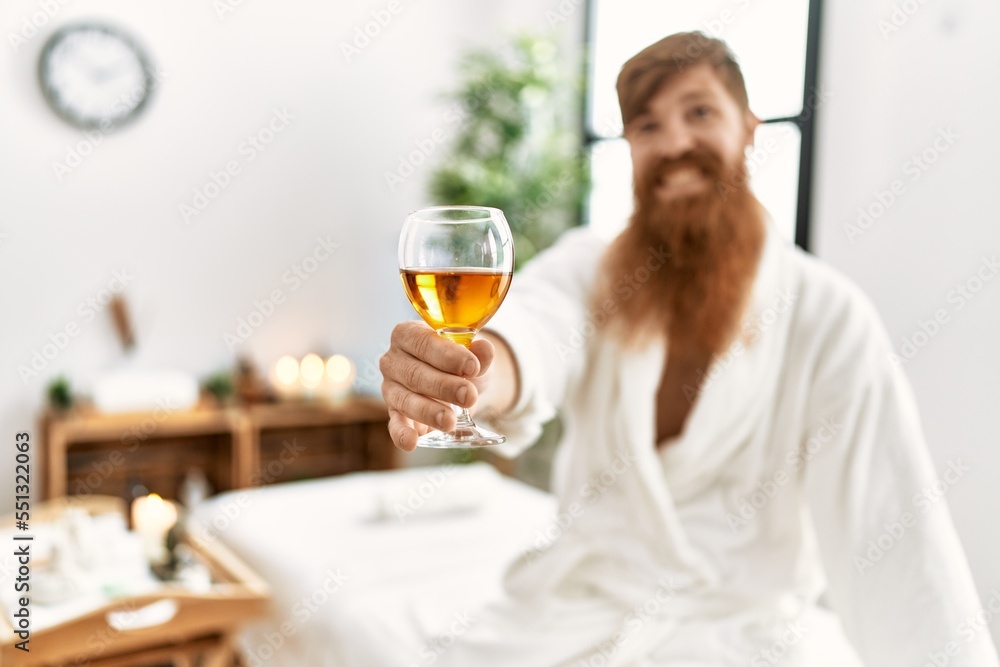 Young redhead man wearing bathrobe drinking champagne at beauty center