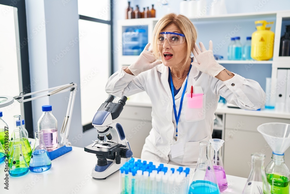Middle age blonde woman working at scientist laboratory trying to hear both hands on ear gesture, curious for gossip. hearing problem, deaf Middle age blonde woman working at scientist laboratory trying to hear both hands on ear gesture, curious for gossip. hearing problem, deaf