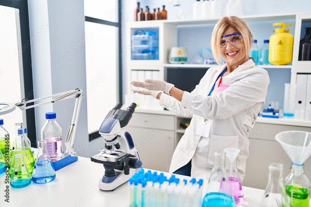 Middle age blonde woman working at scientist laboratory inviting to enter smiling natural with open hand Middle age blonde woman working at scientist laboratory inviting to enter smiling natural with open hand