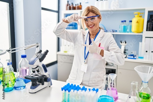 Middle age blonde woman working at scientist laboratory smiling making frame with hands and fingers with happy face. creativity and photography concept.