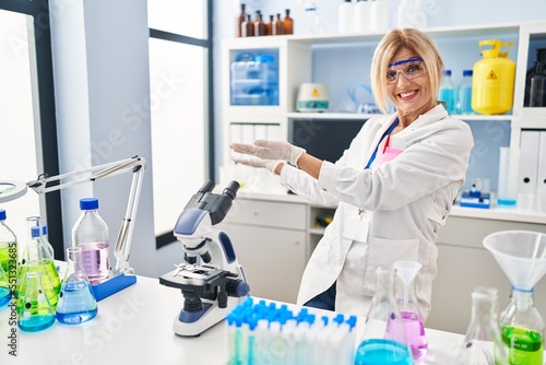 Middle age blonde woman working at scientist laboratory inviting to enter smiling natural with open hand
