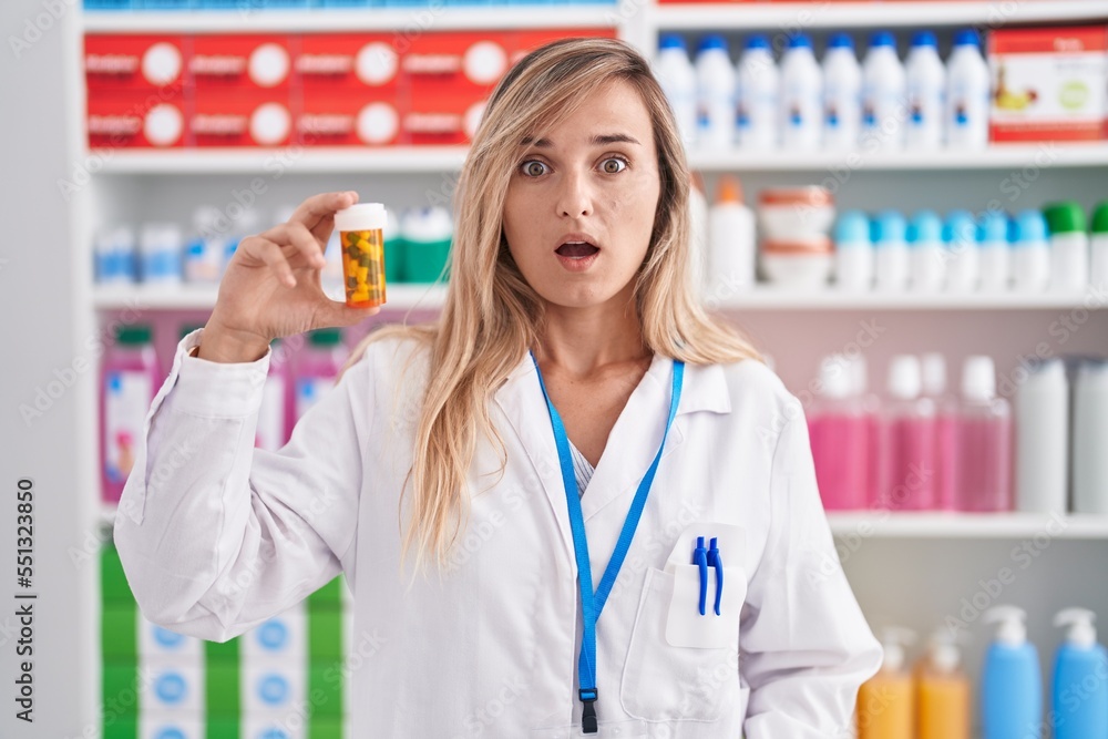 Young blonde woman working at pharmacy drugstore holding pills scared and amazed with open mouth for surprise, disbelief face