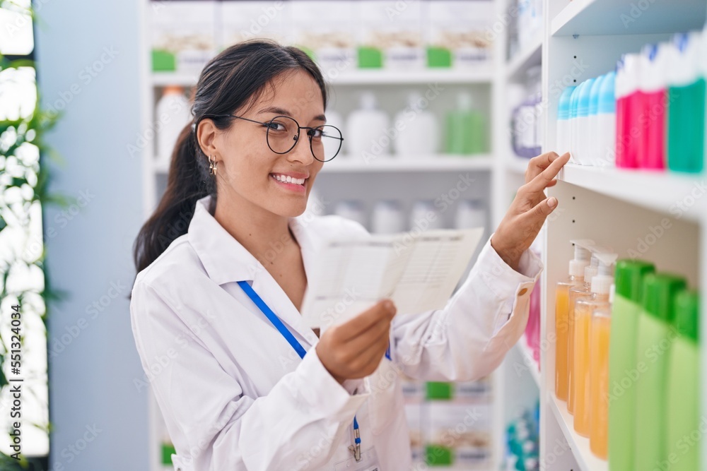 Young beautiful hispanic woman pharmacist reading prescription looking shelving at pharmacy