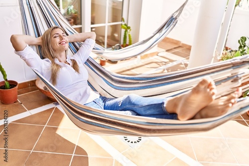 Young blonde woman relaxed with hands on head lying on hammock at terrace.