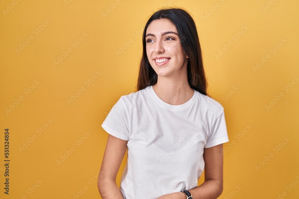 Young beautiful woman standing over yellow background looking away to side with smile on face, natural expression. laughing confident. Young beautiful woman standing over yellow background looking away to side with smile on face, natural expression. laughing confident.