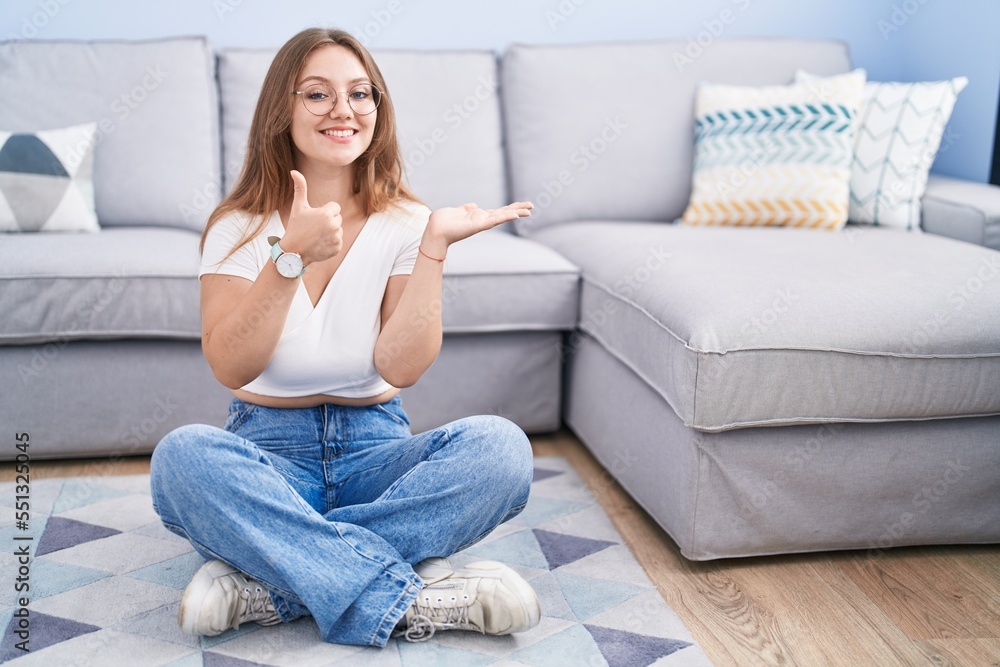 Young caucasian woman sitting on the floor at the living room showing palm hand and doing ok gesture with thumbs up, smiling happy and cheerful