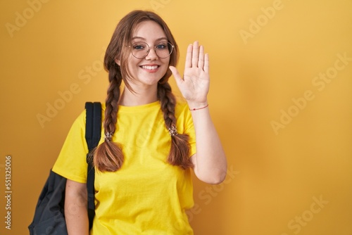 Young caucasian woman wearing student backpack over yellow background waiving saying hello happy and smiling, friendly welcome gesture