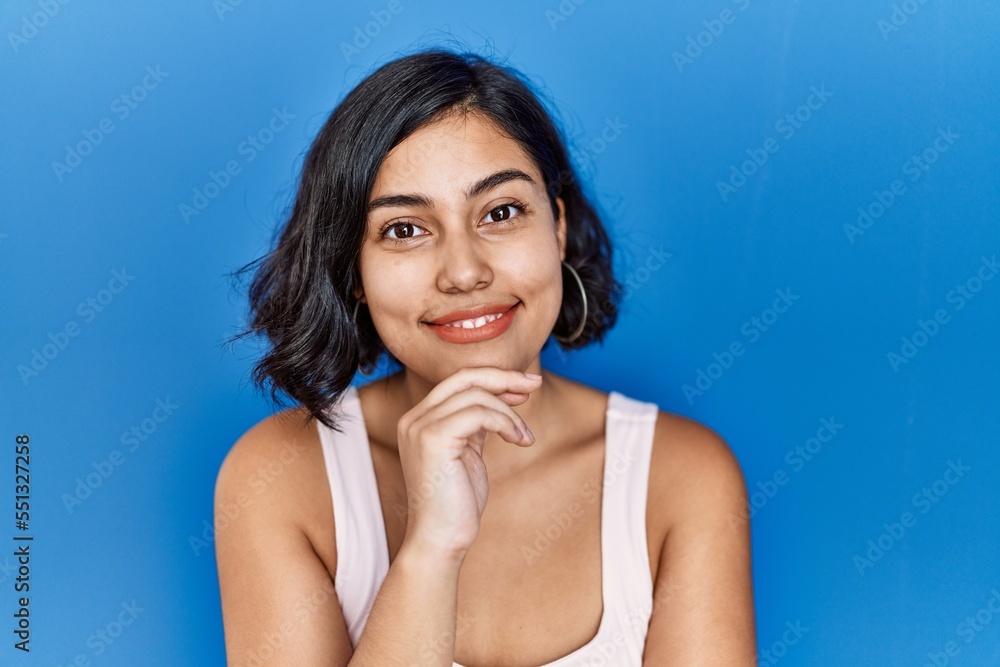 Young hispanic woman standing over blue background looking confident at the camera smiling with crossed arms and hand raised on chin. thinking positive. Young hispanic woman standing over blue background looking confident at the camera smiling with crossed arms and hand raised on chin. thinking positive.