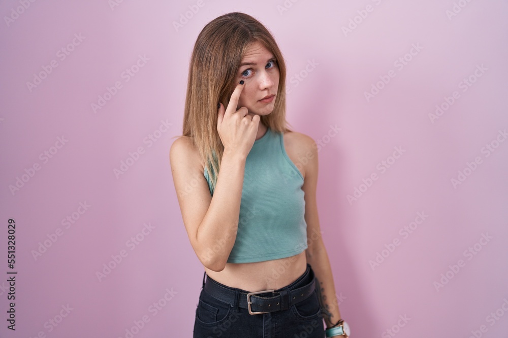 Blonde caucasian woman standing over pink background pointing to the eye watching you gesture, suspicious expression Blonde caucasian woman standing over pink background pointing to the eye watching you gesture, suspicious expression