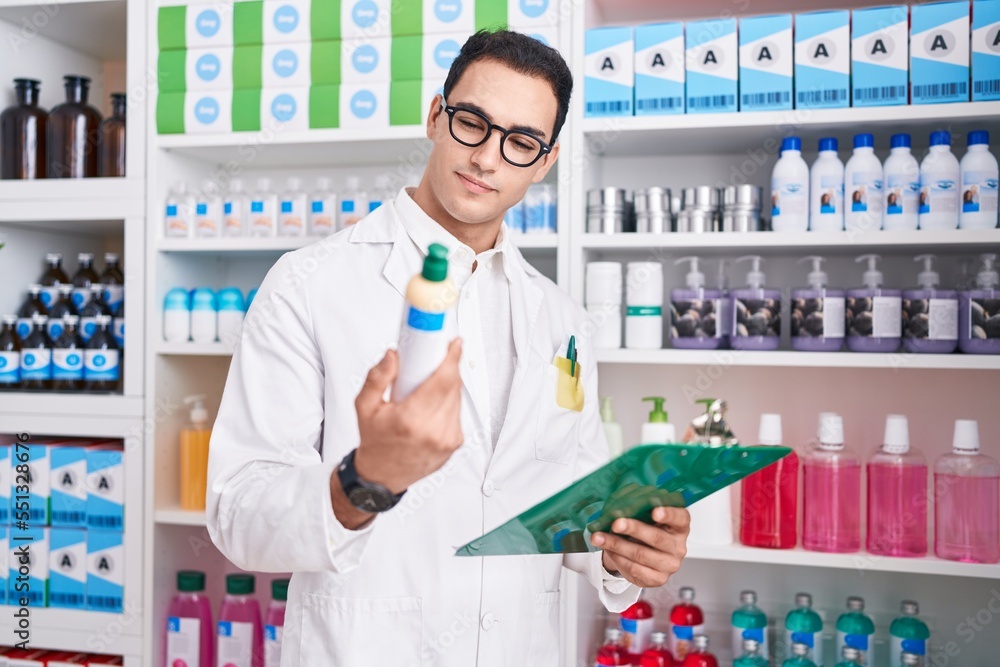 Young hispanic man pharmacist reading document holding shampoo at pharmacy