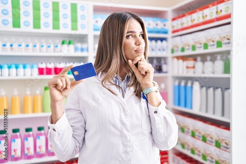 Hispanic young woman working at pharmacy drugstore holding credit card serious face thinking about question with hand on chin, thoughtful about confusing idea