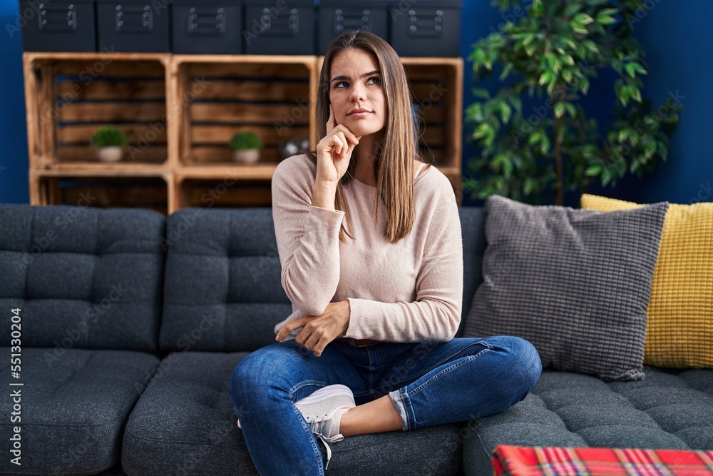 Beautiful woman sitting on the sofa at home serious face thinking about question with hand on chin, thoughtful about confusing idea