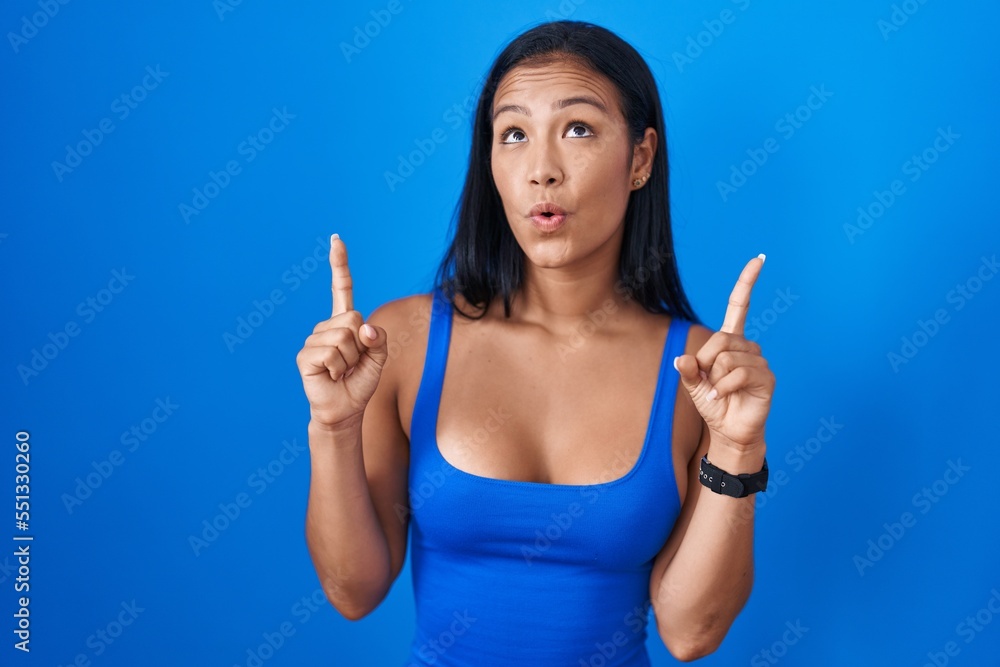 Hispanic woman standing over blue background amazed and surprised looking up and pointing with fingers and raised arms. Hispanic woman standing over blue background amazed and surprised looking up and pointing with fingers and raised arms.