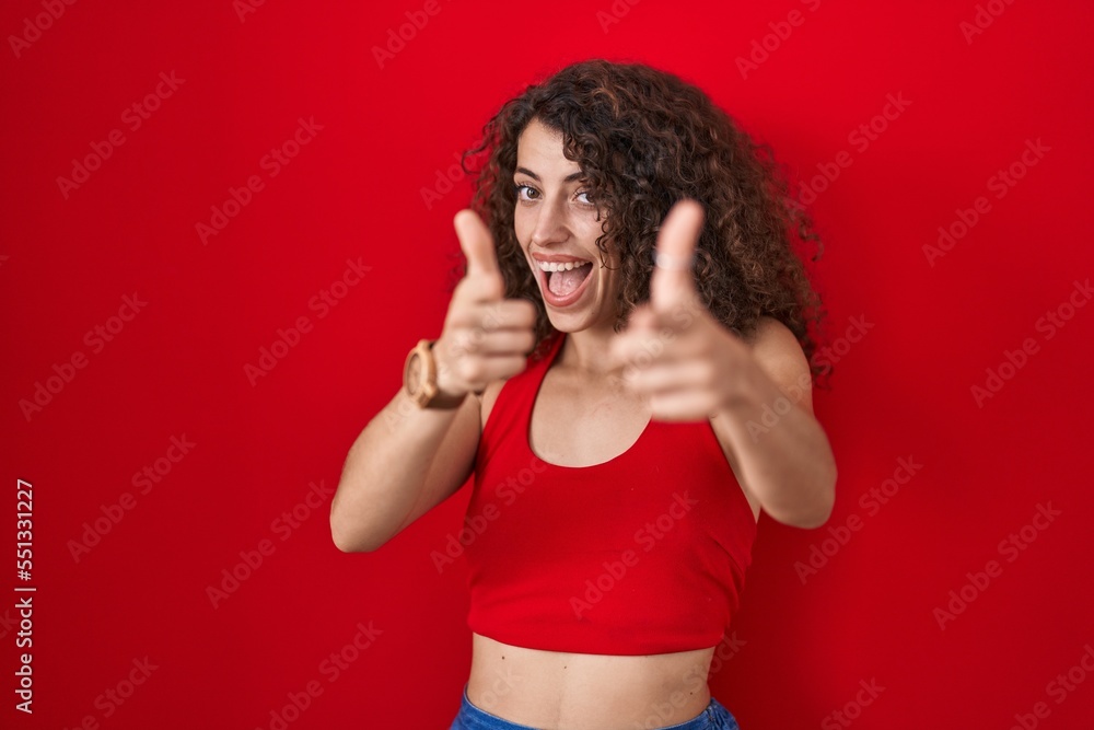Hispanic woman with curly hair standing over red background pointing fingers to camera with happy and funny face. good energy and vibes. Hispanic woman with curly hair standing over red background pointing fingers to camera with happy and funny face. good energy and vibes.