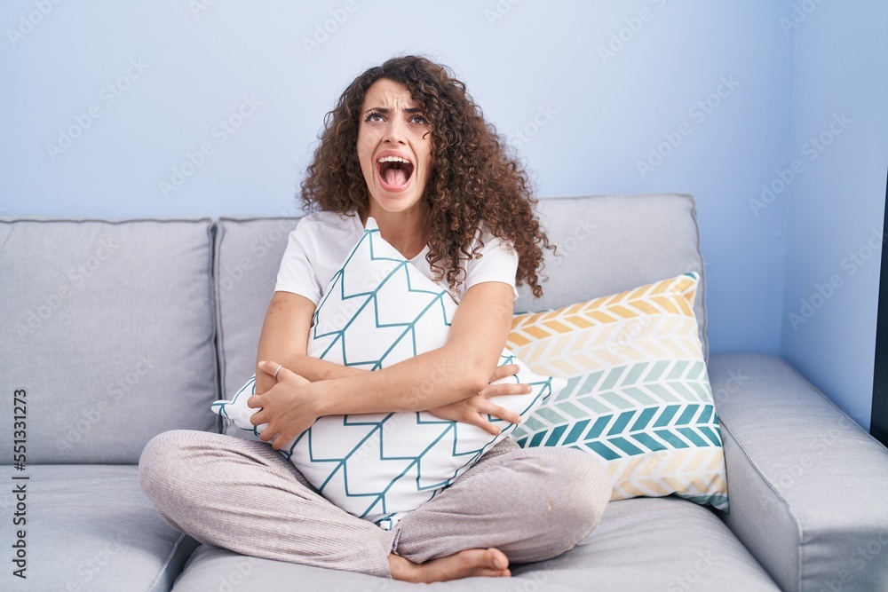 Hispanic woman with curly hair sitting on the sofa at home angry and mad screaming frustrated and furious, shouting with anger looking up. Hispanic woman with curly hair sitting on the sofa at home angry and mad screaming frustrated and furious, shouting with anger looking up.