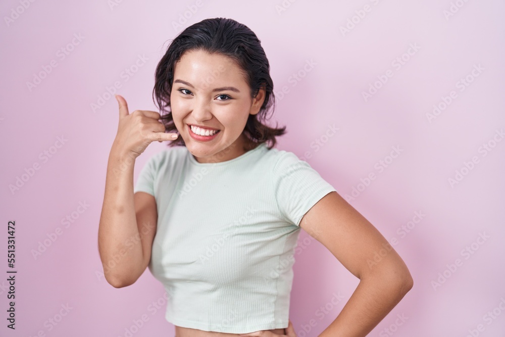 Hispanic young woman standing over pink background smiling doing phone gesture with hand and fingers like talking on the telephone. communicating concepts. Hispanic young woman standing over pink background smiling doing phone gesture with hand and fingers like talking on the telephone. communicating concepts.