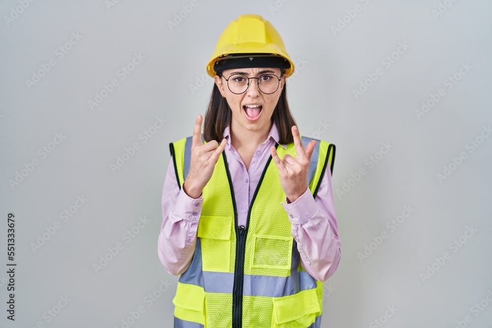 Hispanic girl wearing builder uniform and hardhat shouting with crazy expression doing rock symbol with hands up. music star. heavy concept. Hispanic girl wearing builder uniform and hardhat shouting with crazy expression doing rock symbol with hands up. music star. heavy concept.