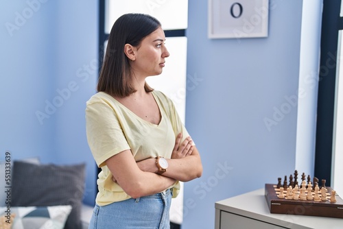 Young hispanic woman standing with serious expression and arms crossed gesture at home