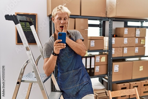 Картината върху платно Young blond man using smartphone working at storehouse afraid and shocked, surpr