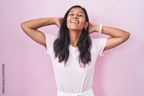 Young hispanic woman standing over pink background relaxing and stretching  arms and hands behind head and neck smiling happy