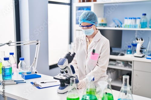 Young blonde woman wearing scientist uniform and medical mask using microscope at laboratory