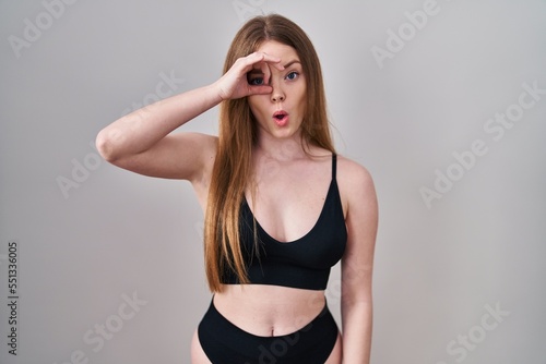 Young caucasian woman wearing lingerie doing ok gesture shocked with surprised face, eye looking through fingers. unbelieving expression.