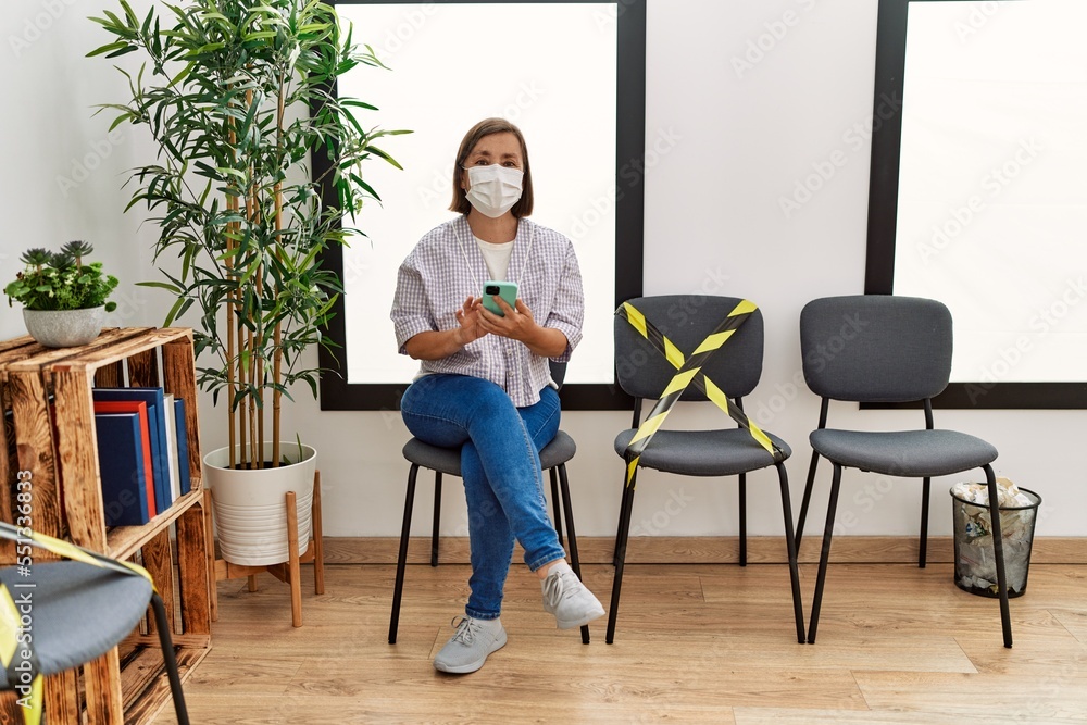 Middle age hispanic woman wearing safety mask using smartphone at waiting room