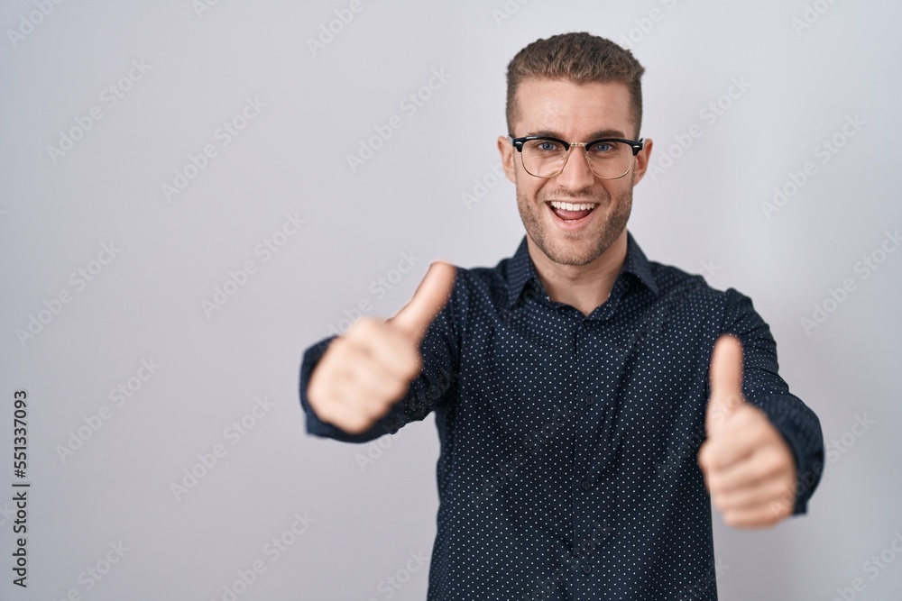 Young caucasian man standing over isolated background approving doing positive gesture with hand, thumbs up smiling and happy for success. winner gesture. Young caucasian man standing over isolated background approving doing positive gesture with hand, thumbs up smiling and happy for success. winner gesture.