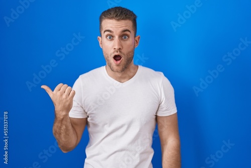 Young caucasian man standing over blue background surprised pointing with hand finger to the side, open mouth amazed expression.