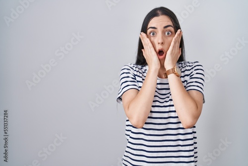 Young brunette woman wearing striped t shirt afraid and shocked, surprise and amazed expression with hands on face