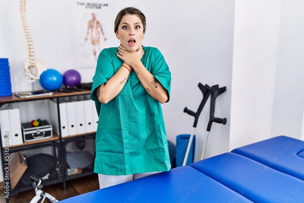 Young hispanic woman wearing physiotherapist uniform standing at clinic shouting and suffocate because painful strangle. health problem. asphyxiate and suicide concept. Young hispanic woman wearing physiotherapist uniform standing at clinic shouting and suffocate because painful strangle. health problem. asphyxiate and suicide concept.