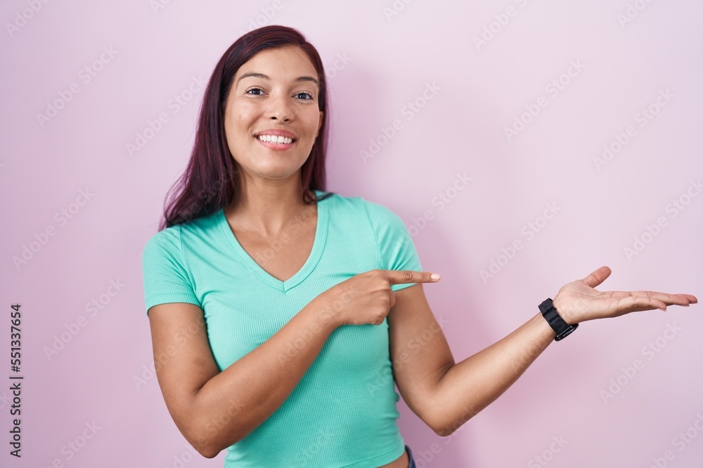 Young hispanic woman standing over pink background amazed and smiling to the camera while presenting with hand and pointing with finger. Young hispanic woman standing over pink background amazed and smiling to the camera while presenting with hand and pointing with finger.